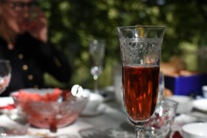 Das Bild zeigt im Vordergrund ein Weinglas zur Hälfte mit Rotwein oder Traubensaft gefüllt, im Hintergrund verschwommen ist ein gedeckter Tisch zu sehen, auf dem eine Glasschale mit Wassermelonenstücken steht.