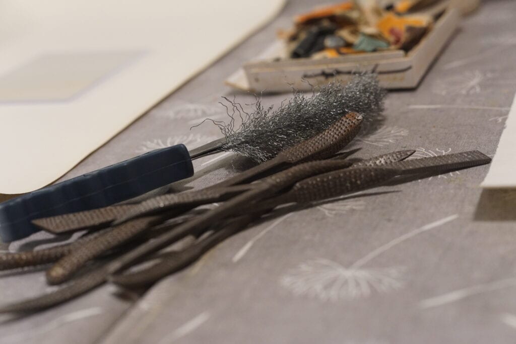 auf einem Arbeitstisch liegen Pfeilen und eine Drahtbürste. Im Hintergrund unscharf weitere Arbeitsutensilien.