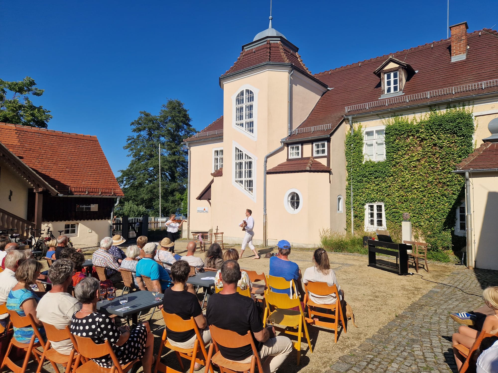 Das Bild zeigt den Innenhof des Kollwitz Hauses. In ihm sitzen Gäste auf Holzstühlen. Sie werden von hinten gezeigt. Sie blicken auf einen Theaterdarsteller.