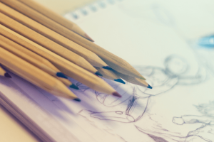 auf einem Zeichenblock liegen hölzerne Buntstifte. Die erste Seite des Blocks zeigt mit Bleistift skizzierte spielende Kinder.