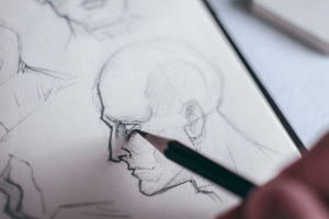 eine Hand hält einen Bleistift. Sie skizziert ein Gesicht. Der Rand des Gesichtes ist sehr deutlich gezeichnet. Im Gesicht Skizzenstriche. Auge, Nase, Mund Ohr sind fertig, Haare fehlen.